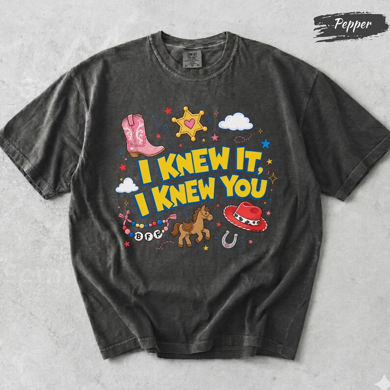 May include: A charcoal gray short-sleeve t-shirt featuring the yellow text I KNEW IT, I KNEW YOU. The graphic includes a pink cowboy boot, a yellow sheriff star, a brown horse, a red cowboy hat, a horseshoe, and a beaded necklace with BFF text.