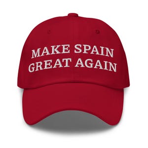 May include: A red baseball cap featuring the white embroidered text MAKE SPAIN GREAT AGAIN across the front. The cap has a structured crown, a curved brim, and a button at the top center, showcasing a classic six-panel design with eyelets.