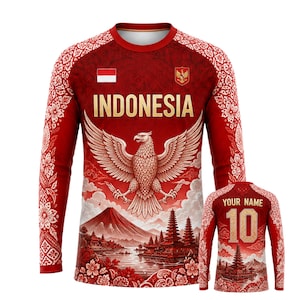 Può includere: Una maglia a maniche lunghe rossa con la scritta INDONESIA in oro, un grande emblema dell'aquila e un grafico paesaggistico con una montagna e templi. Le maniche hanno motivi floreali bianchi. Il retro mostra il testo YOUR NAME e il numero 10.