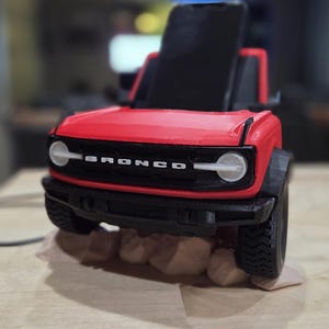 May include: A red and black toy Bronco SUV with the word "BRONCO" across the front. The toy has a black top and sits on a small platform with rocks. The tires are black and the headlights are white.