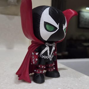May include: A stylized vinyl figure of the character Spawn, featuring a black suit with white markings, a large flowing red cape, silver spiked gauntlets and boots, and a silver chain belt with skull accents. The figure has large green eyes.
