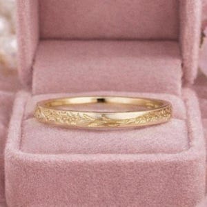 Könnte beinhalten: Ein goldener Ring mit eingraviertem floralem Muster liegt in einer offenen, rosa Samt-Ringbox. Der Ring steht im Mittelpunkt, vor einem Hintergrund aus rosa Stoff, Perlen und Blumen. Das Design des Rings ist deutlich sichtbar.