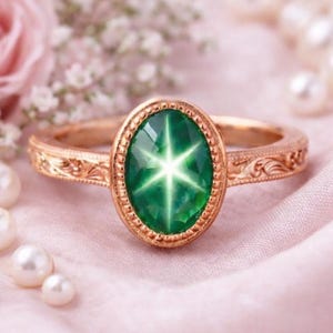 Könnte beinhalten: Ein goldener Ring mit einem ovalen, sternförmigen grünen Edelstein. Der Ring hat ein verziertes, graviertes Band und liegt vor einem rosa Hintergrund mit Perlen und Blumen. Der Edelstein hat ein sechszackiges Sternmuster.