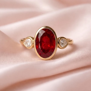 Könnte beinhalten: Ein goldener Ring mit einem großen, oval geschliffenen, tiefroten Edelstein im Zentrum, flankiert von zwei kleineren, birnenförmigen, klaren Edelsteinen. Der Ring ist vor einem weichen, rosa Hintergrund platziert, der das elegante Design des Schmucks hervorhebt.