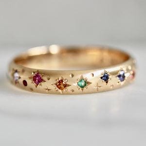 Könnte beinhalten: Ein goldfarbener Ring mit einer Reihe farbiger Edelsteine, die in Stern- und Punktmustern angeordnet sind. Der Ring hat eine glatte, polierte Oberfläche und besteht wahrscheinlich aus Metall. Die Edelsteine sind in verschiedenen Farben gehalten, darunter Rot, Blau, Grün und Orange.