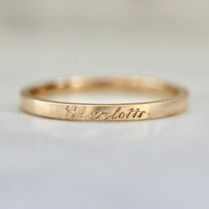 Puede incluir: Un anillo dorado con el nombre "Charlotte" grabado en la banda. El anillo tiene un diseño simple y elegante y un acabado pulido. El anillo es probablemente de oro.