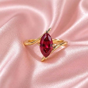 Könnte beinhalten: Ein goldener Ring mit einem tiefroten, marquiseförmigen Edelstein. Das Band des Rings umschließt den Stein und erzeugt ein einzigartiges und elegantes Design. Der Ring liegt auf einem weichen, rosa Satinhintergrund, der seine Farbe und seinen Glanz hervorhebt.
