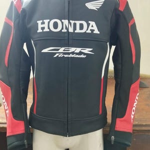 Könnte beinhalten: Eine schwarze Motorradjacke mit roten Akzenten an den Ärmeln und Schultern. Die Vorderseite zeigt den weißen Honda-Schriftzug, ein CBR Fireblade-Logo und ein kleines Honda-Flügel-Emblem auf der Brust. Die Jacke hat einen durchgehenden Frontreißverschluss.