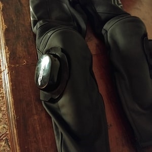 May include: Black leather motorcycle pants featuring integrated knee protection with removable black plastic sliders. The sliders are marked with the white RST logo. The pants include accordion-style stretch panels above the knees for flexibility.