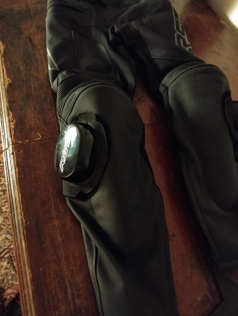 May include: Black leather motorcycle pants featuring integrated knee protection with removable black plastic sliders. The sliders are marked with the white RST logo. The pants include accordion-style stretch panels above the knees for flexibility.