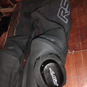 May include: Black motorcycle pants featuring leather construction with accordion stretch panels at the knees. The pants include removable black knee sliders with the white RST logo and a large grey RST logo printed along the upper thigh area.