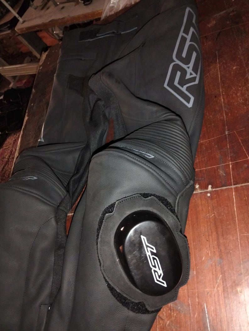 May include: Black motorcycle pants featuring leather construction with accordion stretch panels at the knees. The pants include removable black knee sliders with the white RST logo and a large grey RST logo printed along the upper thigh area.