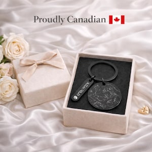 May include: A black metal keyring featuring a circular charm with a white star map engraving and a rectangular tag engraved with the coordinates 43°N 79°25'W, presented in a gift box with a ribbon, accompanied by the text Proudly Canadian.