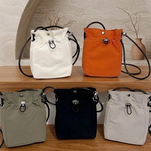 May include: Five bucket bags in white, orange, olive green, black, and light gray. Each bag features a drawstring closure with a silver-toned toggle, a snap button at the top center, and thin black shoulder straps attached to silver metal hardware.