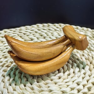 Puede incluir: Una escultura de madera tallada con forma de racimo de plátanos. El objeto presenta un acabado de veta de madera de color marrón claro suave con una forma curva realista, que representa tres plátanos individuales unidos en una base de tallo.