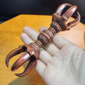 Könnte beinhalten: Ein kupferfarbener Vajra aus Metall mit einem zentralen Griff, der mit komplexen floralen und geometrischen Gravuren versehen ist. Das Objekt hat symmetrische Zinken an beiden Enden, die sich nach innen zu einer Spitze krümmen.