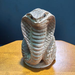 May include: A hand-carved wooden sculpture of a cobra snake in an upright, alert position with its hood flared. The natural light brown wood grain is visible throughout the piece, featuring detailed scale textures on the hood and body.