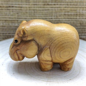 May include: A small, hand-carved wooden figurine of a hippopotamus. The sculpture features a smooth, polished light brown finish with visible natural wood grain patterns throughout the body, rounded ears, and a dark, circular eye detail.