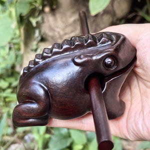 May include: A dark brown wooden frog-shaped percussion instrument featuring a ridged back and a smooth, polished finish. A thin, cylindrical wooden stick is inserted into the side of the frog's mouth, extending outward from the body of the object.
