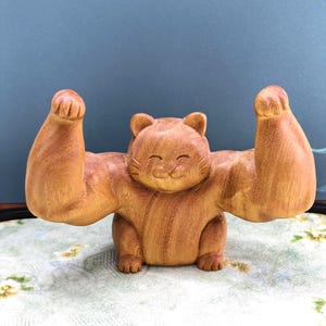 May include: A carved wooden figurine of a cat with an exaggerated muscular physique, featuring large, flexed arms raised in a pose of strength. The cat has a smooth, light brown wood grain finish, pointed ears, and a smiling facial expression.