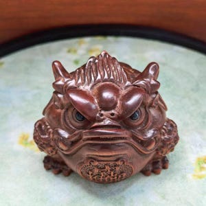 May include: A detailed, brown wooden sculpture of a mythical creature with an angry expression. The figure has large eyes, prominent teeth, and detailed carvings. The sculpture is set against a light green and brown background.