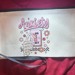 May include: Cream-colored canvas pouch with a black zipper and a striped handle. The pouch features the word "Anxiety" in pink script, a skeleton hand flipping a switch labeled "ON" and "OFF", and floral and star graphics.