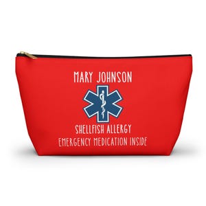 Può includere: Un astuccio rosso con cerniera, cerniera nera e tiretto dorato. L'astuccio presenta il nome "MARY JOHNSON" sopra un simbolo medico blu. Sotto il simbolo, il testo recita "SHELLFISH ALLERGY EMERGENCY MEDICATION INSIDE."
