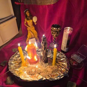 Può includere: Un altare spirituale con una statua, candele e un calice. L'altare è posto su un panno rosso con finiture dorate. Diverse candele accese di varie dimensioni e colori sono disposte su un vassoio rivestito di alluminio con riso e monete.