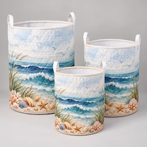 Könnte beinhalten: Ein Set aus drei zylindrischen, gesteppten Stoffkörben mit einer Küstenszene aus blauen Meereswellen, Sandstrand, Strandgras, Muscheln, Seesternen und fliegenden Möwen. Jeder Korb ist mit zwei weißen Stoffgriffen ausgestattet.