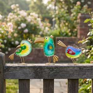 May include: Three colorful metal bird sculptures with gold wire frames and legs. The birds have vibrant blue, green, yellow, and red accents. They are perched on a weathered wooden fence, with a blurred background of flowers.