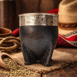 Puede incluir: Una taza de mate de cuero negro con un borde plateado con un diseño en relieve. La taza tiene las palabras "La Gaucha" en la parte delantera. La taza está sobre una superficie de arpillera con una cuchara de madera y hojas de té sueltas.