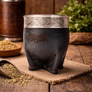 Puede incluir: Una taza de mate de cuero negro con borde plateado y el texto "LA GAUCHITA GIL". La taza está sobre un posavasos de arpillera, con yerba mate suelta y una cuchara de madera cerca.