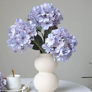 May include: A bouquet of light purple hydrangea flowers in a cream-colored, double-bulb vase. The flowers have multiple layers of petals and are arranged with green leaves. A white teacup with a gold spoon is visible in the background.