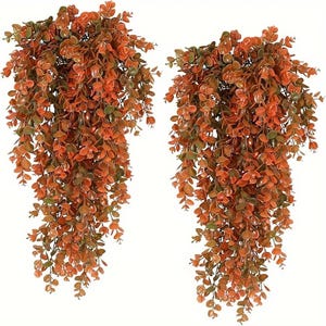 Puede incluir: Dos ramos de plantas artificiales colgantes con tallos en cascada cubiertos de pequeñas hojas redondas de colores otoñales en tonos naranja quemado, óxido y verde apagado. El follaje es denso y texturizado, creando una apariencia llena y colgante.
