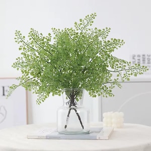 May include: A clear glass vase holds a vibrant arrangement of artificial green fern. The fern has delicate, small leaves and is arranged to create a full, lush appearance. The vase sits on a white surface, suggesting a decorative or home decor theme.
