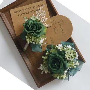 Puede incluir: Dos boutonnieres de rosas verdes y un corsage a juego descansan sobre una bandeja de madera. Los boutonnieres tienen cinta verde y detalles blancos. Una tarjeta con el texto "WONDERFUL THINGS FLOWER STUDIO" está en el fondo.