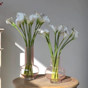 Puede incluir: Dos jarrones de cristal rosa, cada uno con ramos de lirios cala blancos. Los jarrones tienen una base redondeada y un cuello cilíndrico. Las flores tienen tallos verdes, creando una presentación completa y elegante.