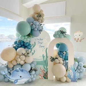 Könnte beinhalten: Eine Partydekoration zum Thema Ozean mit einem Wal-Ausschnitt, Luftballons in Blau-, Weiß- und Beigetönen und einem Hintergrund mit dem Text "Under The Sea". Die Anordnung umfasst einen Bogen und Korallen-Ausschnitte.