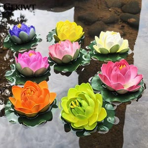 Puede incluir: Nenúfares artificiales flotantes en varios colores, incluyendo azul, amarillo, rosa, blanco, naranja y verde. Cada lirio tiene un diseño detallado de pétalos y descansa sobre una hoja de lirio verde, creando una exhibición vibrante en el agua.