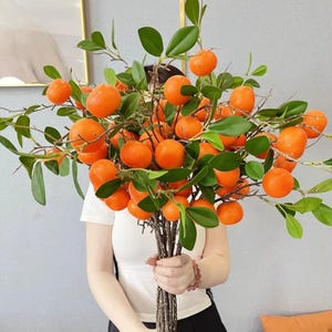 May include: A bunch of artificial orange tangerines with green leaves and brown stems. The bright orange fruit contrasts with the green foliage, creating a visually appealing arrangement. The bouquet is held in front of a person wearing a white top.