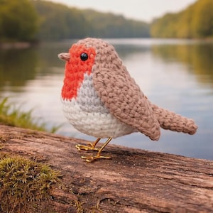 Op de afbeelding: Een handgemaakte gehaakte roodborst met een rode borst, grijs en wit lichaam en bruine vleugels. De vogel heeft zwarte ogen en zit op een boomstam met gouden draadpoten. De achtergrond toont een meer en bomen.