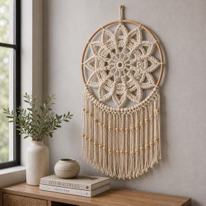 Könnte beinhalten: Ein cremefarbener Makramee-Traumfänger mit einem Holzreifen und Perlenfransen. Das zentrale Design zeigt ein Blumenmuster. Der Traumfänger hängt an einer hellgrauen Wand und verleiht einen Hauch von Bohème-Dekor.