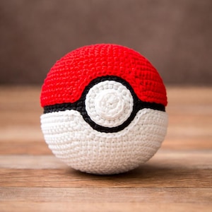 May include: A crocheted Poké Ball in red, white, black, and cream. The top half is red, the bottom half is white, and a black band separates the two. A white circle with a black outline is in the center.