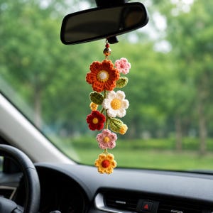 Può includere: Un ciondolo floreale per auto all'uncinetto. Il ciondolo presenta una cascata di fiori nei toni dell'arancione, rosso, bianco, rosa e giallo, con foglie verdi. È appeso allo specchietto retrovisore di un'auto, aggiungendo un tocco decorativo all'interno del veicolo.