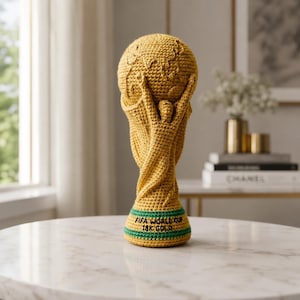 以下が含まれることがあります： 金色の手編みFIFAワールドカップトロフィー。緑色のストライプと「FIFA WORLD CUP 18K GOLD」の文字が入った台座。大理石のテーブルに飾られています。高さは約30cmです。
