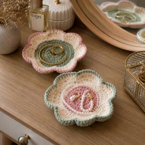 May include: Two handmade, flower-shaped crochet trays in pastel colors. One tray holds a gold ring and bracelet, the other holds a pearl necklace and ring. The trays are on a wooden surface, ideal for jewelry storage.