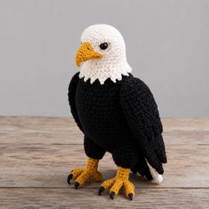 May include: A handmade crochet bald eagle figurine. The eagle has a white head, a yellow beak, and black wings and body. The eagle's talons are yellow with black claws. The figurine is standing on a wooden surface.