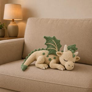 Könnte beinhalten: Ein gehäkeltes Drachenspielzeug in Schlafposition mit cremefarbenem Körper, hellgrünen Flügeln, einem grün bestachelten Schwanz und kleinen Hörnern. Der Drache hat geschlossene Augen und eine strukturierte Garnoberfläche mit detaillierten Maschen.