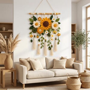 May include: A wall hanging featuring a large central crocheted sunflower surrounded by smaller white and orange crocheted flowers, green leaves, and wooden beads, all suspended from a horizontal wooden rod with cream-colored yarn tassels.