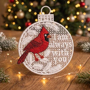 May include: A circular white embroidered ornament featuring a red cardinal bird perched on white pine branches. The text "I am always with you" is stitched in white thread on the right side. The ornament has a loop at the top for hanging.